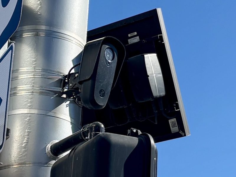 A photo of Flock Safety license plate reader, a black device about the size of a football, mounted on a pole at East 105th Street and Euclid Avenue. A square solar panel is visible behind it.